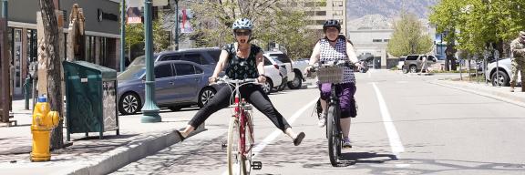 two women riding bikes downtown. One has her feet off the pedals and extended to the side in a playful manner. The other is following behind smiling. 