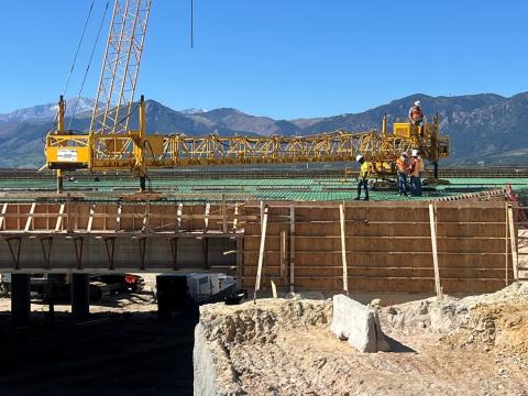 A construction site with a large yellow crane spanning the width of the image, positioned above a partially built concrete structure with wooden formwork. Several workers in safety gear and helmets are seen standing on the platform near the crane. In the background, a mountain range is visible under a clear blue sky.