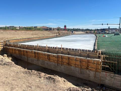 A construction site showing a large, flat concrete surface that appears freshly poured within wooden formwork. The formwork runs along the length of the structure, supported by beams. To the right, green rebar is visible, indicating ongoing reinforcement work. In the background, construction equipment and vehicles are seen, with some buildings and a traffic light visible under a clear blue sky.