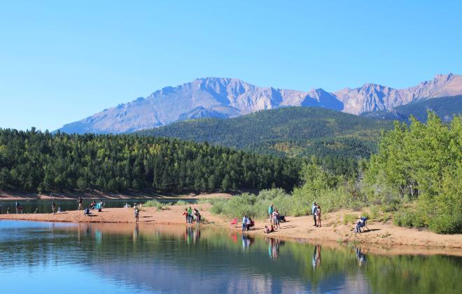 A lush view of participants fishing on a lakeshore on the inaugural Youth Fishing Derby in 2023. Pikes Peak is visible in the background behind smaller mountains covered in trees.