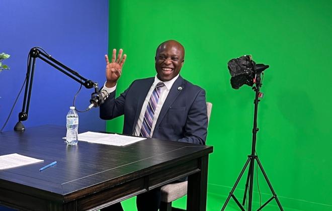 Mayor Yemi smiles at the camera in front of a green screen and holds up four fingers.